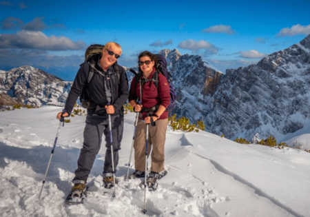 Two smiling hikers with trekking poles on a snowy mountain, with snow-covered peaks and blue sky in the background