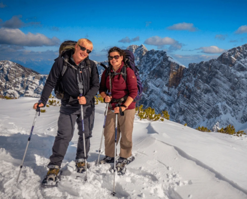 Two smiling hikers with trekking poles on a snowy mountain, with snow-covered peaks and blue sky in the background