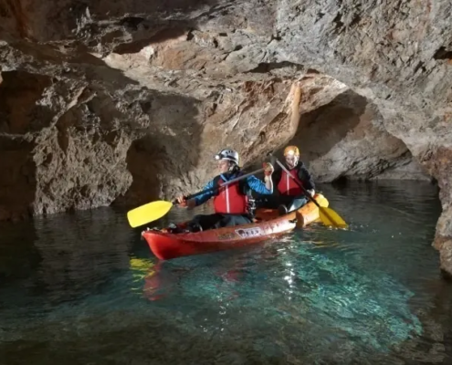 Two people kayaking through an underground cave with clear water and rocky walls, wearing helmets and life jackets.