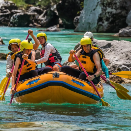 Rafting Slovenia: Group of people wearing helmets and life jackets paddling together on an inflatable raft in a turquoise river, navigating through rocks during a rafting adventure.