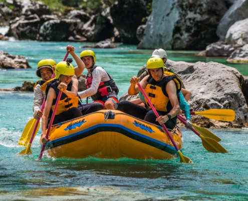 Rafting Slovenia: Group of people wearing helmets and life jackets paddling together on an inflatable raft in a turquoise river, navigating through rocks during a rafting adventure.