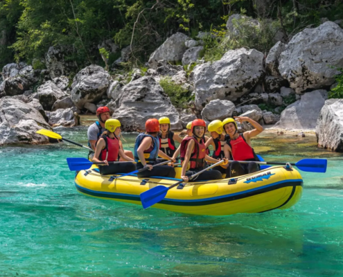 Rafting Slovenia: Group of people on a yellow inflatable raft in a clear turquoise river, wearing helmets and life jackets while smiling and waving, surrounded by rocky riverbanks and lush greenery during a rafting trip.