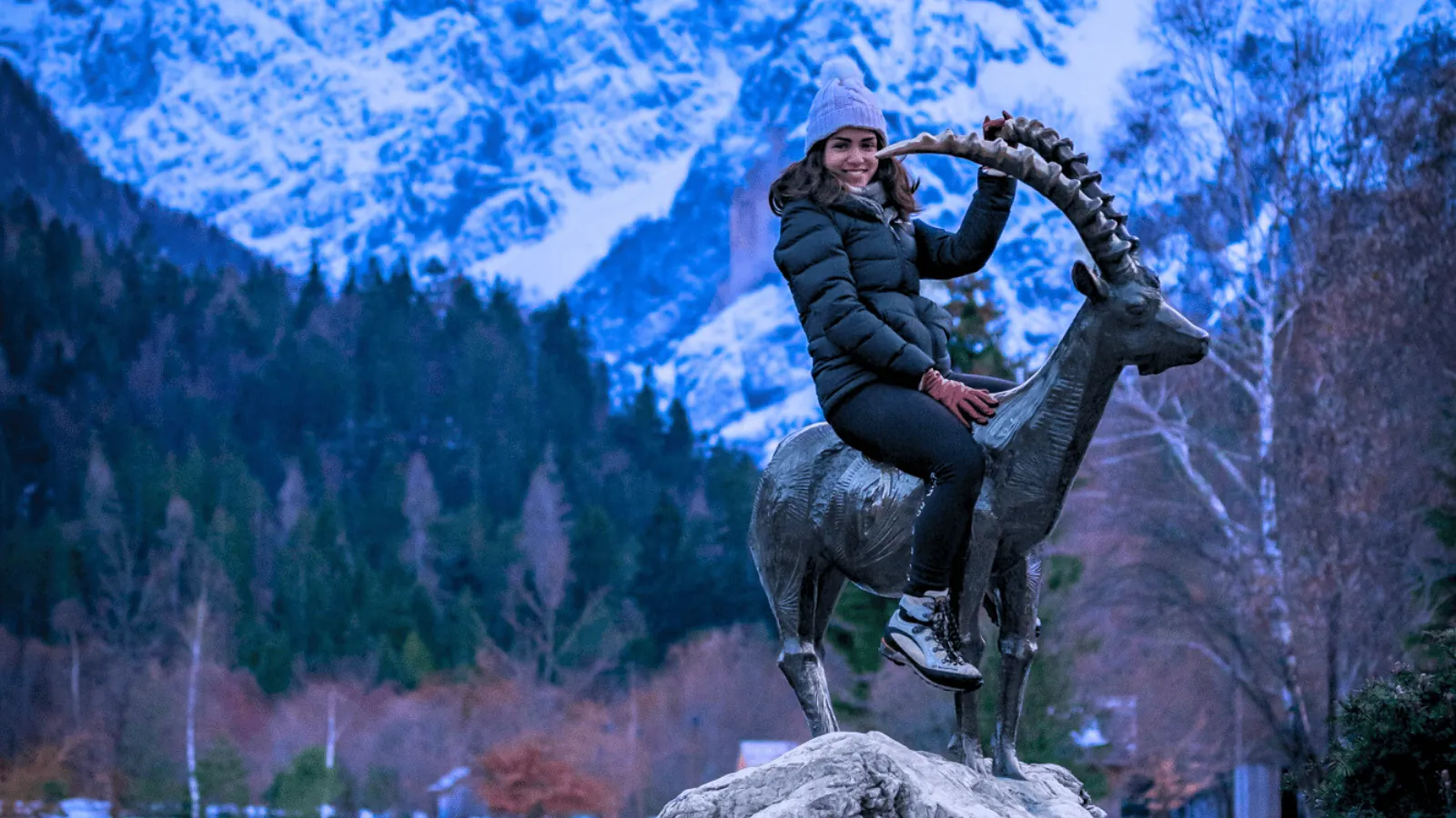 A woman sitting on a statue of an ibex at Jasna Lake in Slovenia, with snow-covered mountains in the background. She is wearing winter clothes, including a puffy jacket and a beanie, smiling against the scenic, wintry landscape during dusk.