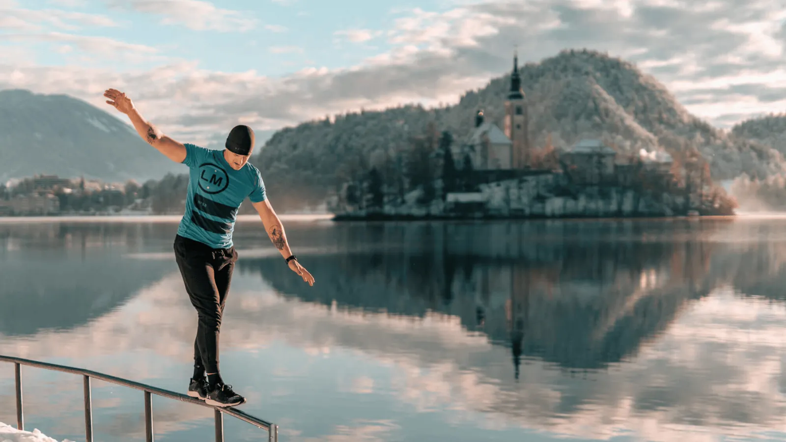 A man walks along a fence in Bled, Slovenia, during winter, wearing shoe sleeves for grip. Snow blankets the ground and surrounding landscape, creating a peaceful, frosty atmosphere.