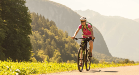 A woman is seen biking on a gravel road on a warm sunny day with picturesque mountains in the background