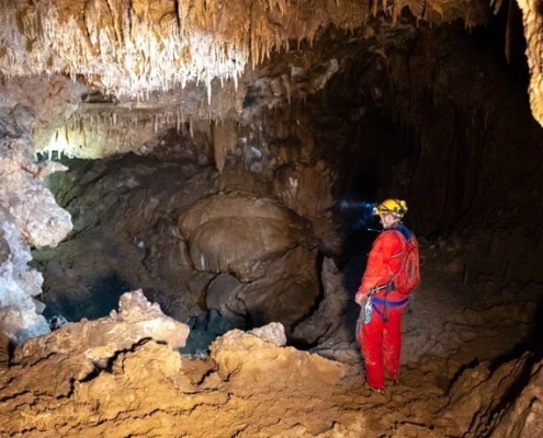 A person wearing red caving gear and a helmet with a headlamp exploring a dimly lit cave, surrounded by stalactites, stalagmites, and rugged rock formations.