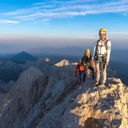 Three climbers in helmets and harnesses roped together, standing on a narrow rocky mountain ridge with expansive alpine views under a blue sky.