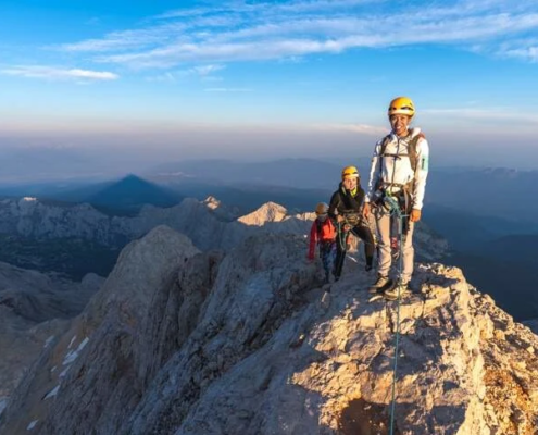 Three climbers in helmets and harnesses roped together, standing on a narrow rocky mountain ridge with expansive alpine views under a blue sky.
