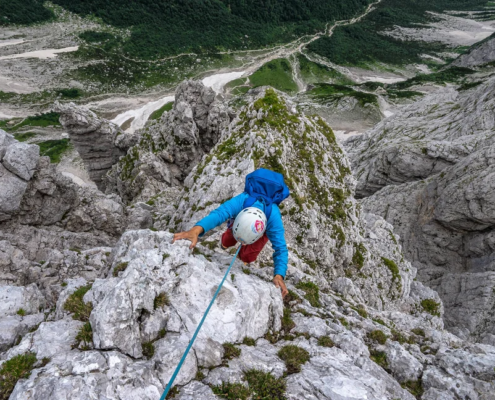 A climber scaling a rugged limestone ridge in Slovenia's Julian Alps, with a rope for safety and lush green valleys stretching out below.