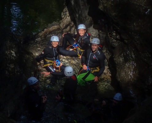A group of adventurers on a night canyoning tour, wearing helmets and wetsuits, gathered in a rocky canyon illuminated by soft light, surrounded by dark shadows