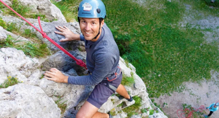 Smiling rock climber is seen from above as he ascends on rocky path in a full safety gear
