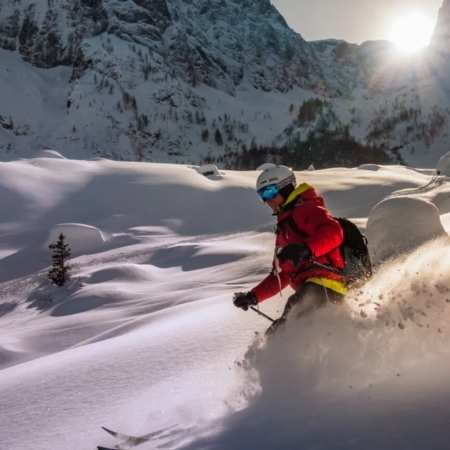 Person skiing through deep powder snow on a sunny day in a mountainous winter landscape, surrounded by snowy peaks and pine trees.