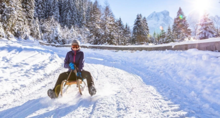 Smiling woman is seen sledding on a snowy road surrounded by trees and mountains