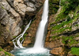 A cascading waterfall flows into a turquoise pool, surrounded by rugged cliffs and lush green vegetation. The scene is vibrant, with moss and trees adding contrast.