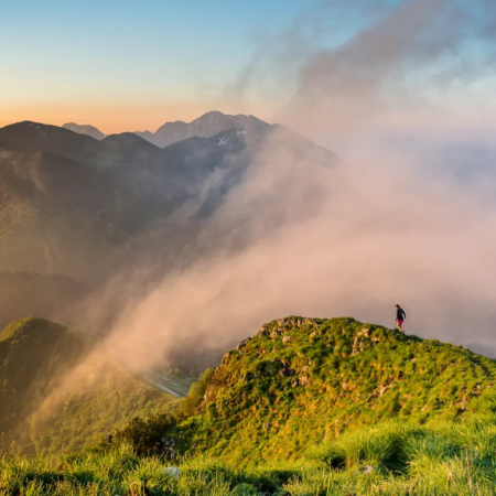 Hiking Slovenia: A lone hiker standing on a misty mountain ridge, surrounded by rolling green hills and soft clouds.