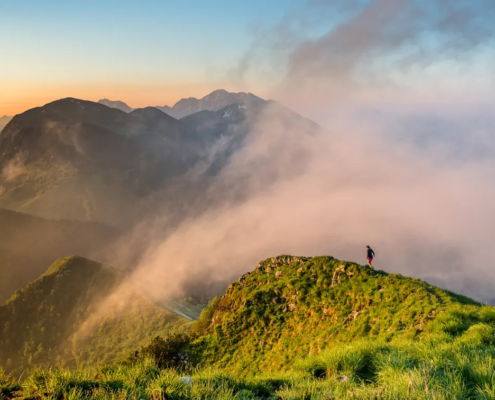Hiking Slovenia: A lone hiker standing on a misty mountain ridge, surrounded by rolling green hills and soft clouds.