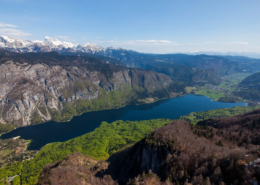 Bled Tours: Panoramic aerial view of a deep blue alpine lake surrounded by forested hills and snow-capped mountains under a clear blue sky.