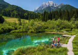 Bled Tours: A group of friends raising hands at the emerald green Zelenci Lake with lush meadows and mountains creating a lively natural backdrop.