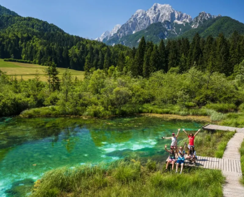 Bled Tours: A group of friends raising hands at the emerald green Zelenci Lake with lush meadows and mountains creating a lively natural backdrop.