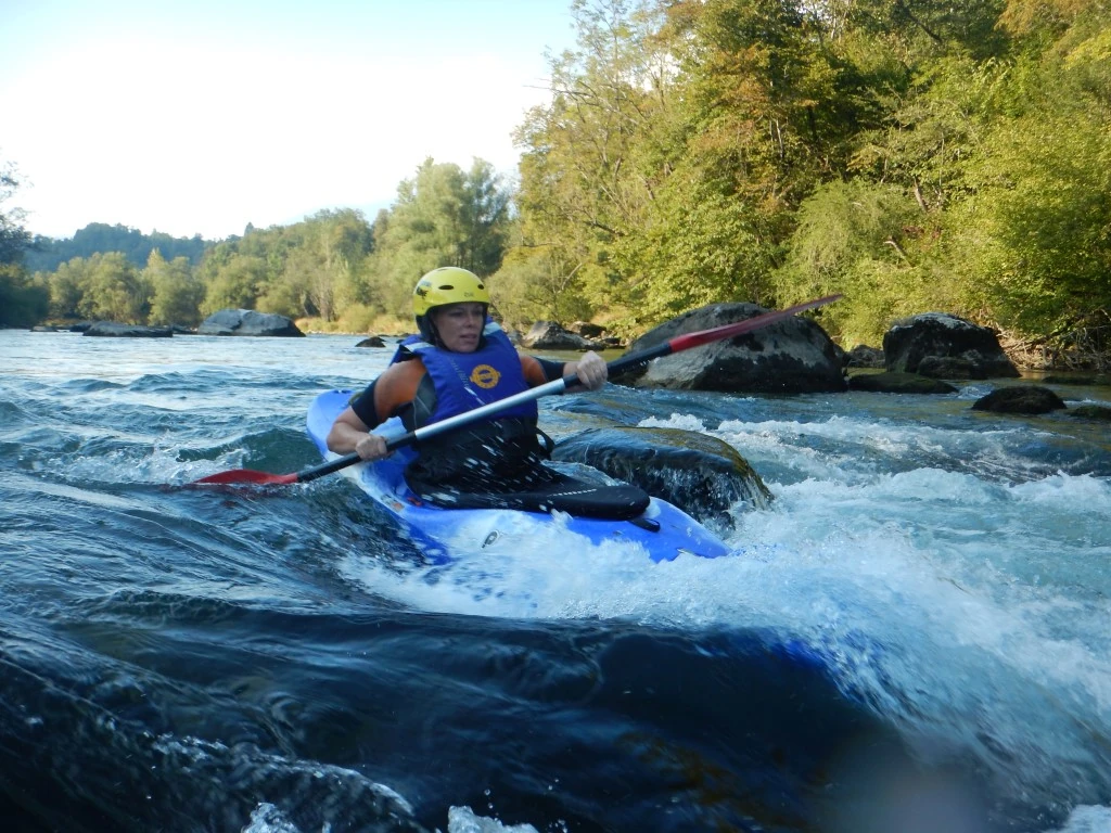 Kayaking Kayaking Slovenia: Kayaker paddling through fast river currents under sunlight, surrounded by forested banks.
