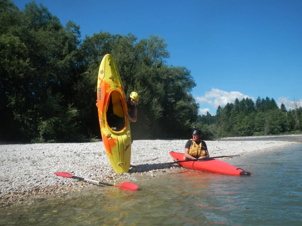Kayaking Kayaking Slovenia: Person flips a kayak upright on a gravel beach beside calm blue river water and pine trees.