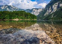 Slovenia Tours: Crystal-clear waters of Lake Bohinj reflect surrounding mountains and forests under a bright blue sky with scattered clouds.