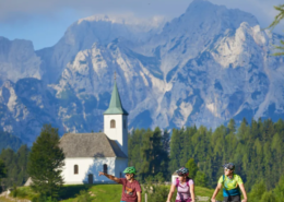 Slovenia Tours: Cyclists riding on a mountain road near a white chapel with forested slopes and towering jagged peaks in the background.