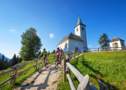 Slovenia Tours: Group of people riding downhill with a small white church in a background on a sunny day, surrounded by grass and mountains.