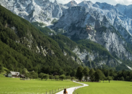 Slovenia Tours: Person walking on a paved path through a green valley with alpine mountains and a partly cloudy blue sky in the background.