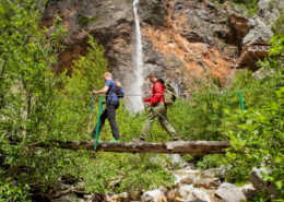 Bled Tours: Two hikers crossing a rustic wooden bridge over a stream with a tall waterfall and dense forest in the alpine landscape behind them.