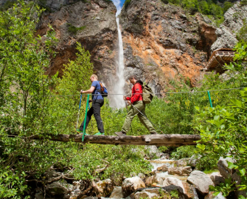 Bled Tours: Two hikers crossing a rustic wooden bridge over a stream with a tall waterfall and dense forest in the alpine landscape behind them.