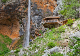 Slovenia Tours: Narrow trail passing by a tall waterfall cascading down a rocky cliff with a wooden viewing platform nestled among lush greenery.