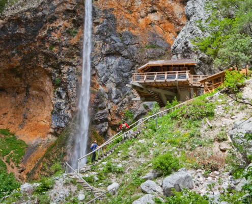 Slovenia Tours: Narrow trail passing by a tall waterfall cascading down a rocky cliff with a wooden viewing platform nestled among lush greenery.