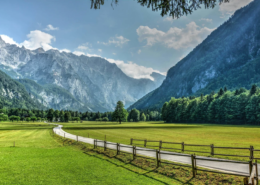 Slovenia Tours: Scenic view of Logar Valley with a winding road, green meadows, and towering alpine mountains under a partly cloudy sky.