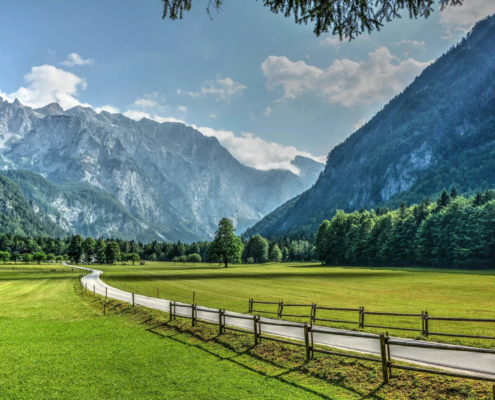 Slovenia Tours: Scenic view of Logar Valley with a winding road, green meadows, and towering alpine mountains under a partly cloudy sky.