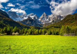 Slovenia Tours: The Špik mountain range rises sharply in the distance, surrounded by grassy meadows and a dramatic sky filled with fluffy clouds.