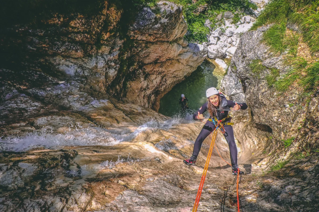 Woman abseiling down a sunlit waterfall in Fratarica canyon, giving a thumbs up, with lush green surroundings and a pool below