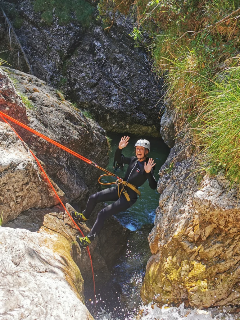 Canyoner lowering themselves into a narrow rocky chute using ropes and a bright orange harness.