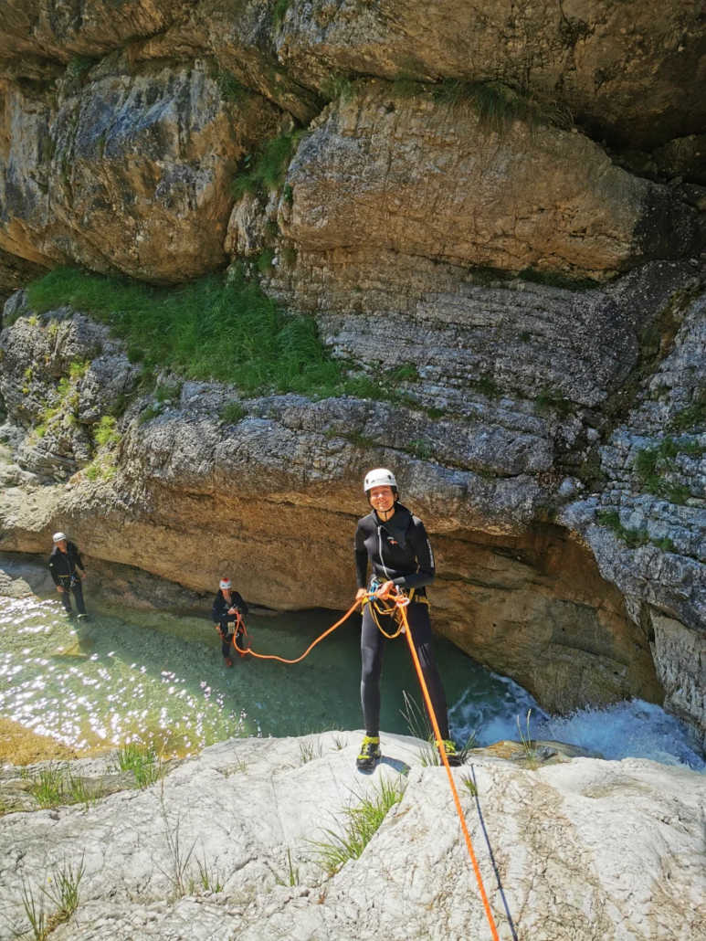 Adventurer preparing to rappel down a rocky drop with crystal-clear water below in a sunlit canyon.