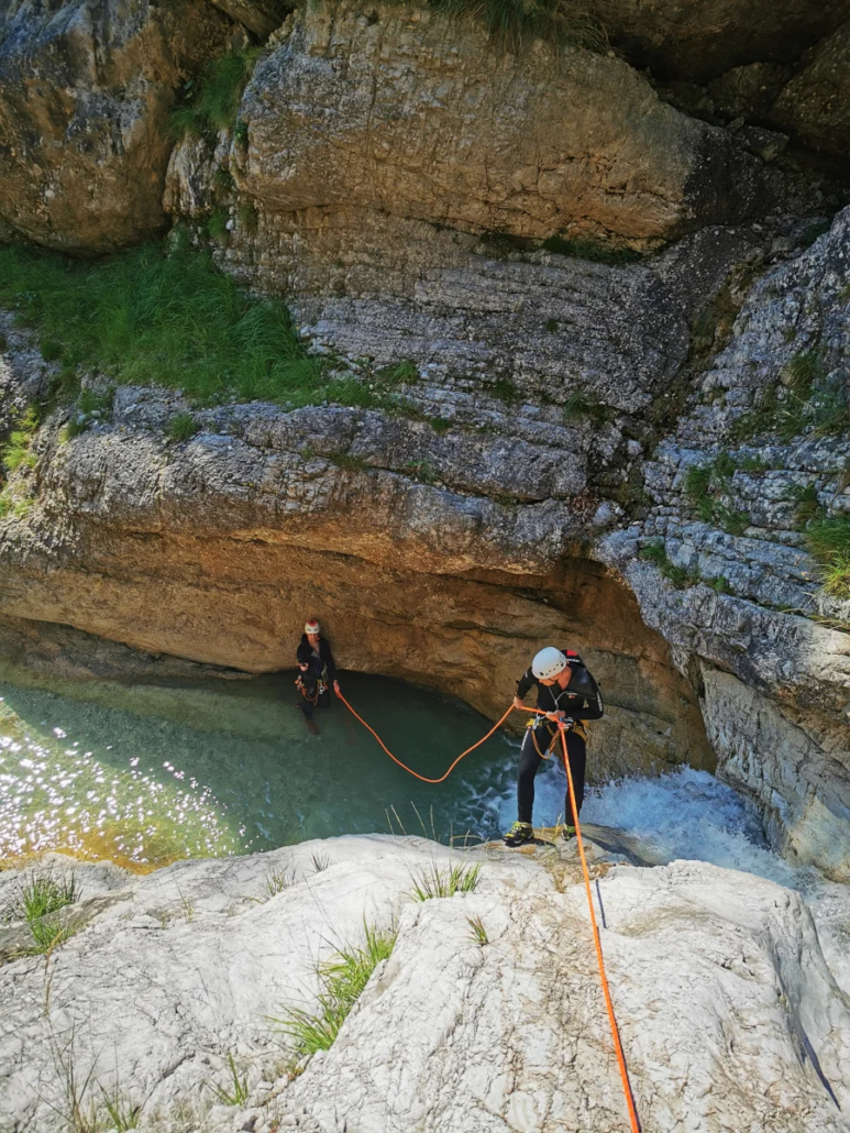 Person abseiling down into a canyon pool with layered rock formations surrounding the descent path.
