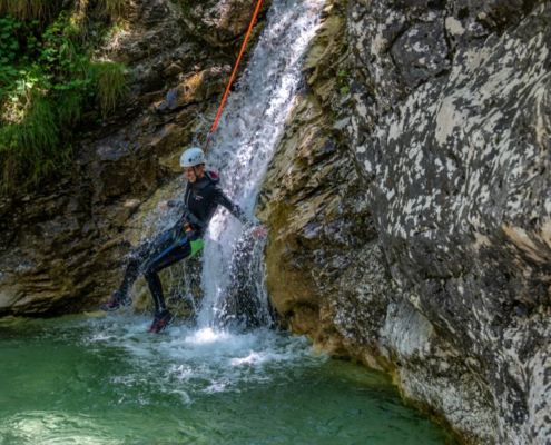 Canyoner descending a small waterfall on a rope, landing in a clear green pool surrounded by mossy rock and forest.
