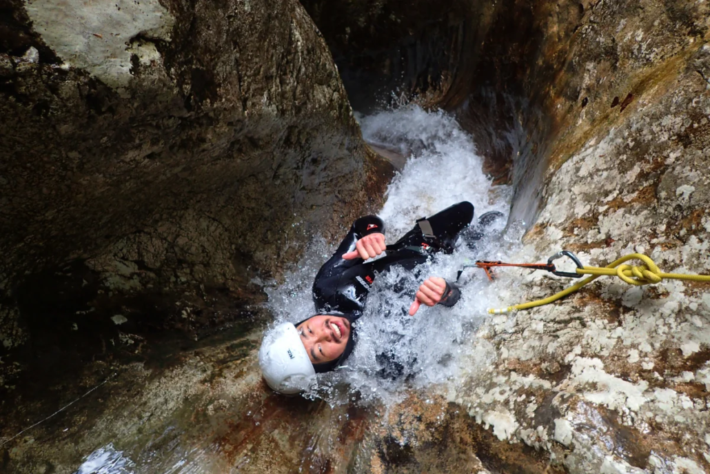 Canyoner sliding down a steep, wet rock chute into fast-moving water, helmet on and arms up.