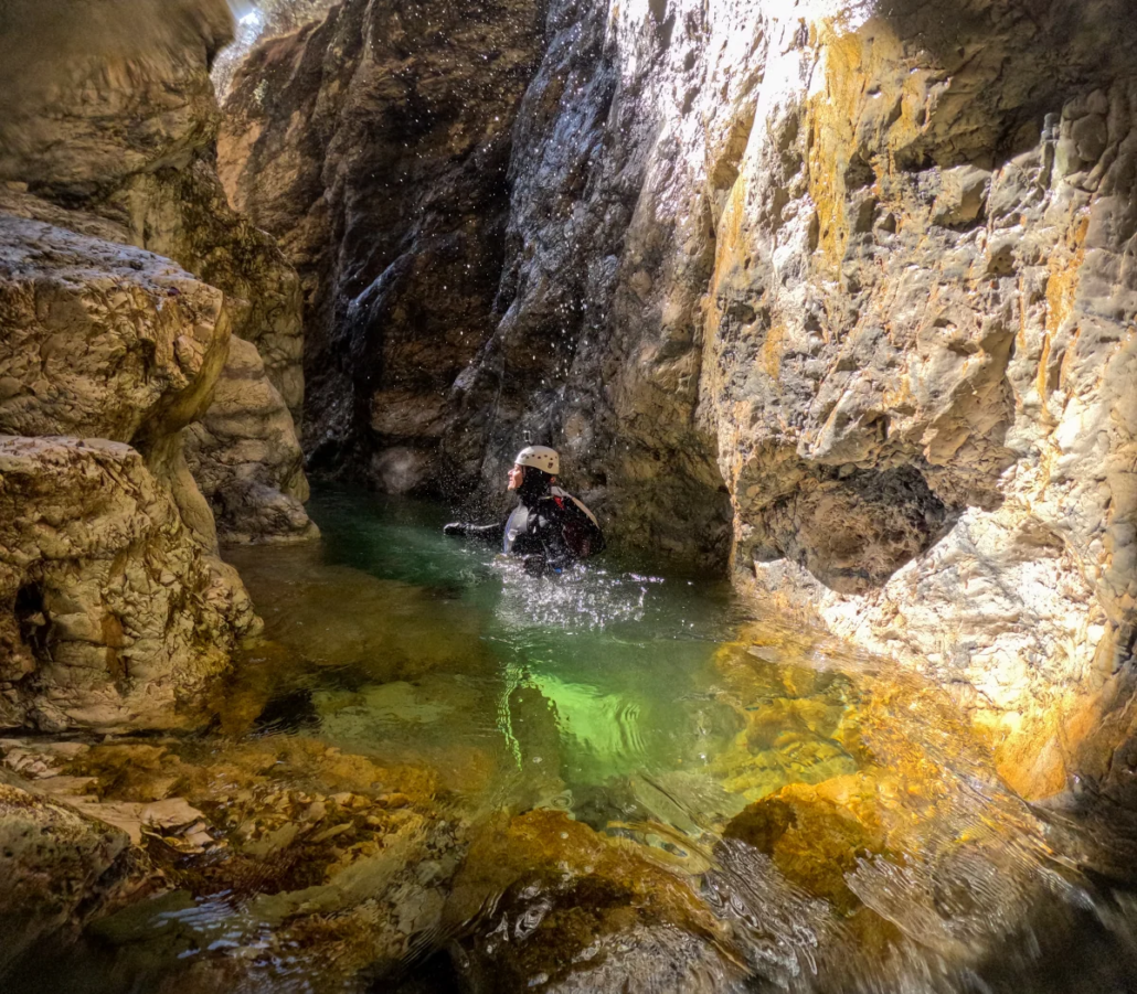 Person floating in a calm green pool, surrounded by tall rock walls with sunlight shining through.
