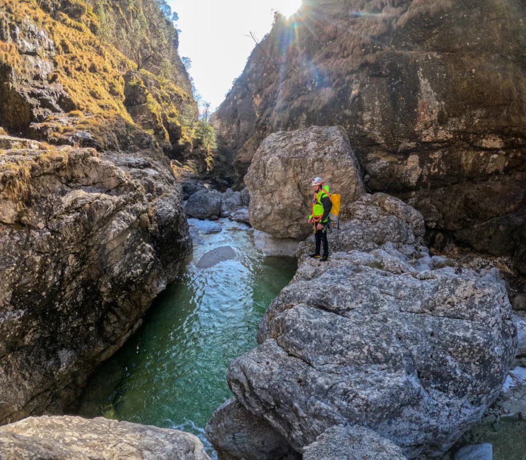 Canyoner walking along rocky ledges beside emerald-green water in a sunlit canyon.