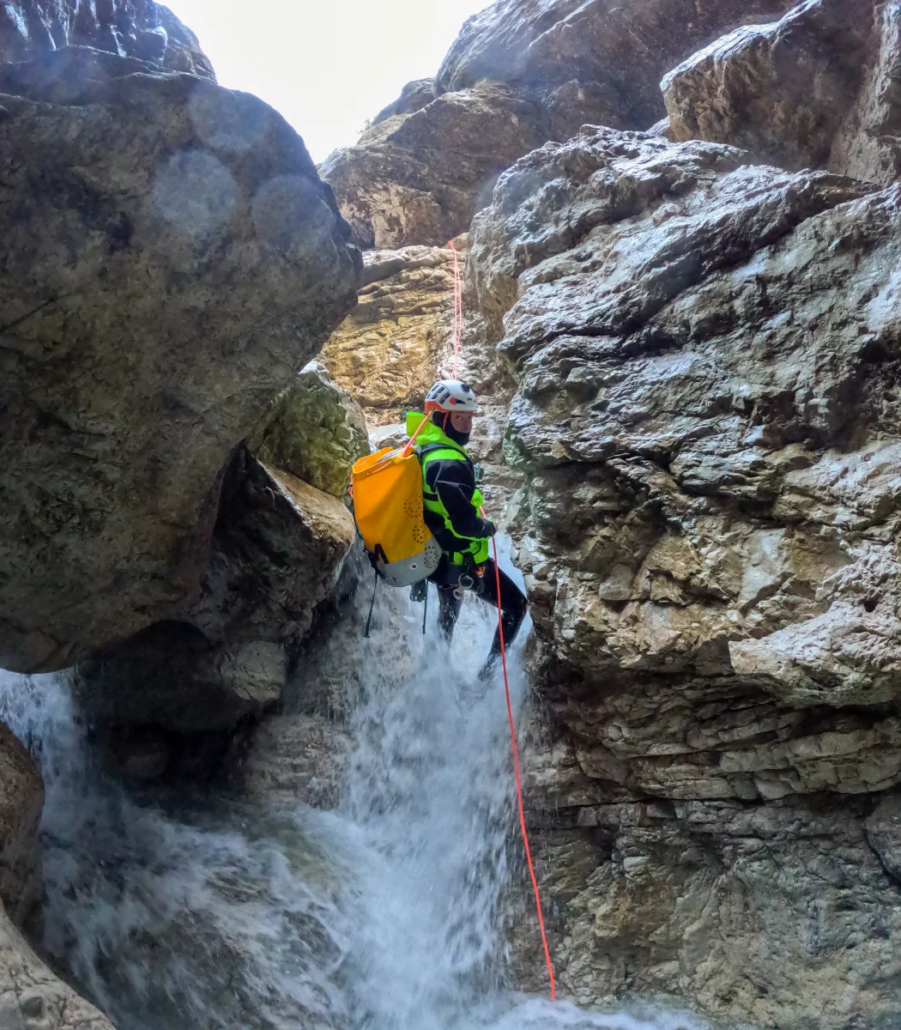 Adventurer rappelling down a steep waterfall between narrow rock formations with a yellow pack.