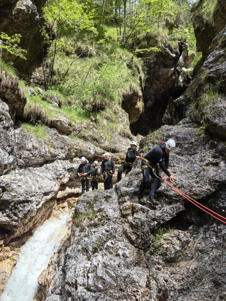 Line of canyoners walking along a rocky path with ropes in a green forested canyon area.
