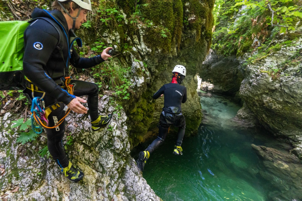 Person jumps into a natural pool in a narrow canyon ledge above a turquoise stream, wearing helmets and wetsuits.