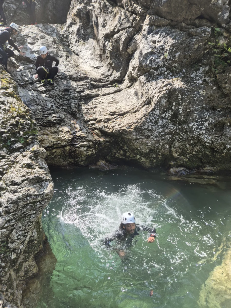 Canyoner landing in a deep green pool as another watches from the rock above in a narrow canyon.