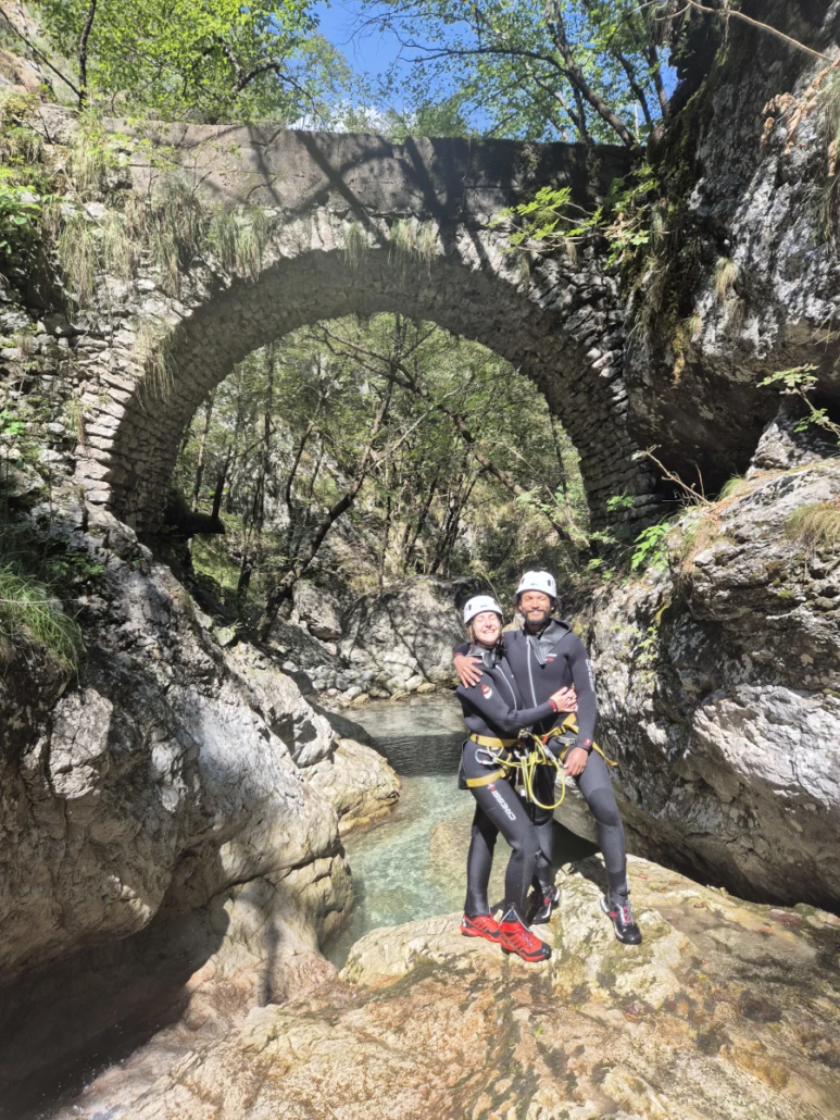 Two canyoners in wetsuits standing beneath a stone arch bridge over a shallow canyon stream.