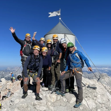 Group of climbers smiling and raising their arms next to the Aljaž Stolp on the summit of Mount Triglav
